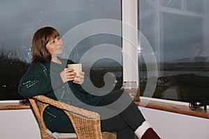 Woman sitting in a cozy wicker chair by the wide window on the veranda, drinking tea, concept of rest and relaxation