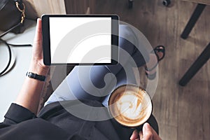 A woman sitting cross legged and holding black tablet pc with blank white desktop screen while drinking coffee in cafe