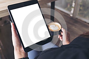 A woman sitting cross legged and holding black tablet pc with blank white desktop screen while drinking coffee in cafe