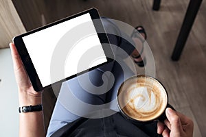 A woman sitting cross legged and holding black tablet pc with blank white desktop screen while drinking coffee in cafe