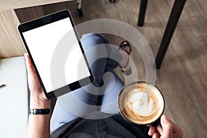 A woman sitting cross legged and holding black tablet pc with blank white desktop screen while drinking coffee in cafe