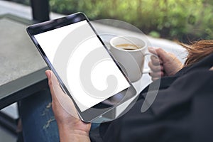 A woman sitting cross legged and holding black tablet pc with blank white desktop screen while drinking coffee in cafe