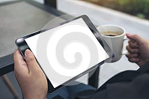 A woman sitting cross legged and holding black tablet pc with blank white desktop screen while drinking coffee in cafe