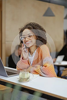 A woman is sitting at a desk with a laptop, talking on a cell phone and smiling