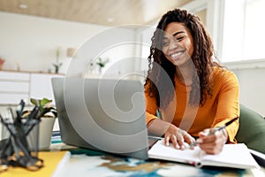 Woman sitting at desk, using computer and writing in notebook