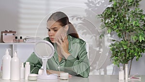 A woman sitting at a dressing table in front of a mirror scrutinizes her face. A young woman with problematic skin