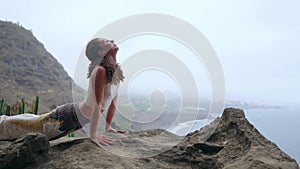 The woman sitting on the edge of a cliff in the pose of the dog with views of the ocean, breathe in the sea air during a