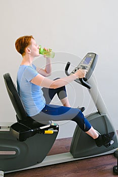 Woman sitting on exercise bike in the gym and drinking water from a sports bottle