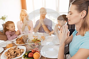 A woman is sitting at a festive table and praying. Against the background, her family sits and also pray