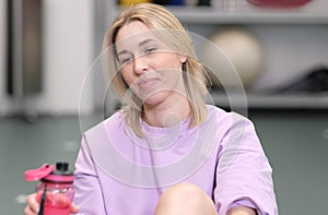 woman sitting on a floor in a gym and getting rest on workout. blond woman holding water bottle in fitness club.