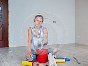 A woman sitting on the floor mixes glue in a red bucket for wallpapering in the apartment.