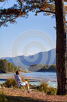 A woman is sitting on a folding chair on the bank of a mountain river