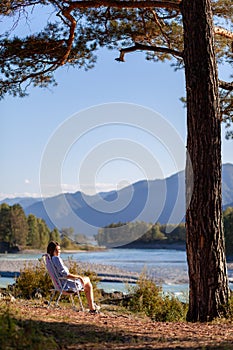 A woman is sitting on a folding chair on the bank of a mountain river