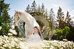 Woman sitting in a forest spring sun lit meadow full of daisy flowers, white Arabian horse next to her