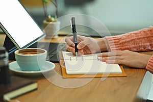 Woman sitting in front of computer tablet and writing information on notebook.
