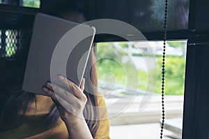 A woman sitting by glass window looking , using and holding tablet pc cover her face