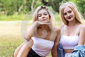 Woman sitting on green grass with her friends while looking at camera. Joyful two teenagers girl sitting relaxed