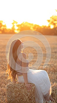 Woman sitting on hay stack walking in summer evening, beautiful romantic girl with long hair outdoors in field at sunset