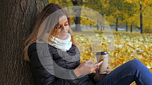 Woman Sitting With Her Back To The Tree In Yellow Autumn Leaves, Uses Smartphone