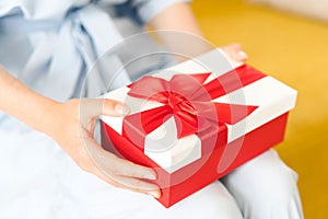 Woman sitting and holding infront of her beautiful giftbox with red bow in hands. Festive red and white present box