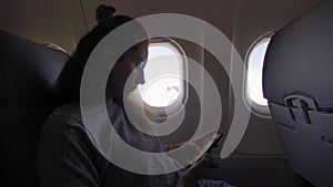 Woman is sitting inside a plane, looking in porthole and texting messages in her phone.