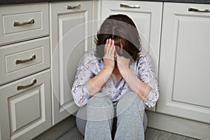 Woman is sitting on kitchen floor covering her face with her hands. Depression, grief or frustration