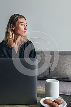 Woman sitting with laptop on the couch in the morning, working at home, watching morning news over a cup of coffee