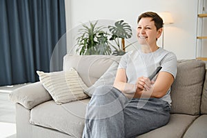 Woman sitting in living room holding remote control smiling.