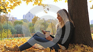 Woman Sitting Near Tree In Yellow Fall Leaves, Uses Apps And Drinking Coffee