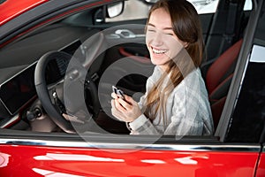 Woman sitting in new red car holding key rejoicing her purchase in dealership