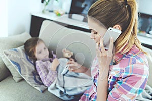 A woman is sitting next to a little girl who is sick. She holds a thermometer, which the girl`s temperature measured.