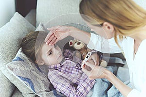 A woman is sitting next to a little girl who is sick. She holds a thermometer, which the girl`s temperature measured.