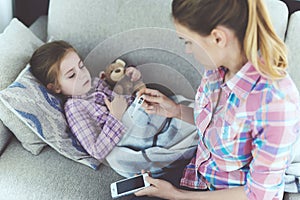 A woman is sitting next to a little girl who is sick. She holds a thermometer, which the girl`s temperature measured.