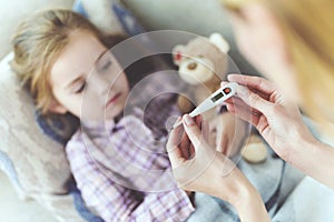 A woman is sitting next to a little girl who is sick. She holds a thermometer, which the girl`s temperature measured.