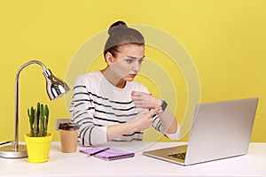 Woman sitting in office and pointing to watch, looking at laptop screen, exhausted of overtime work.