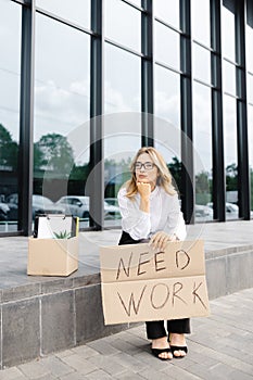 Woman sitting outdoor with cardboard banner with the slogan Need work next to box of stuff. Beautiful female protester at strike