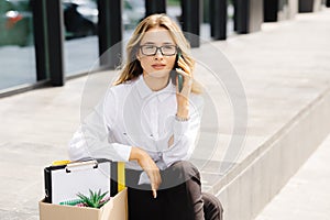 Woman sitting outdoor with cardboard banner with the slogan Need work next to box of stuff and talking to the phone. Beautiful