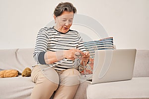 Woman sitting on sofa and looking at the laptop screen while knitting