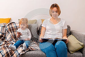 A woman is sitting on the sofa and reading a book. A small child is sitting next to him and playing on the phone. Family