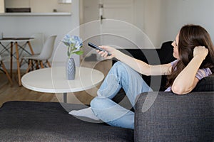 A woman is sitting on a sofa a watching TV at home. Girl is using remote control