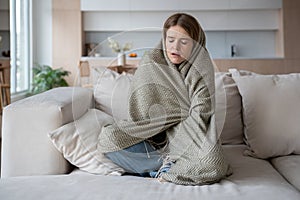 Woman sitting on sofa wrapped in plaid, having shivering, temperature, ill with epidemic flu, virus