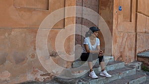 Woman sitting on steps in front of doorway with cap and sneakers looking to her left