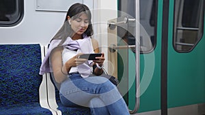 Woman sitting in a subway car, wearing headphones, watching video on a phone