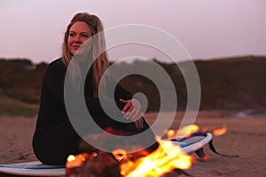 Woman Sitting On Surfboard By Camp Fire On Beach As Sun Sets Behind Her