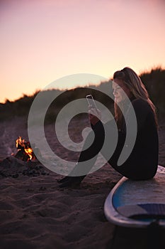 Woman Sitting On Surfboard By Camp Fire On Beach Using Mobile Phone As Sun Sets Behind Her