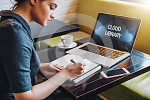 Woman sitting at table in cafe in front of laptop computer with inscription on monitor-cloud library and making notes in notebook