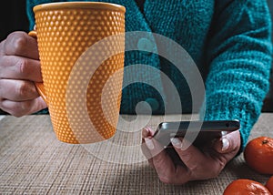 A woman is sitting at a table holding a cup of tea and a mobile phone