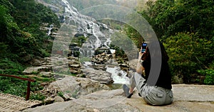 Woman sitting and taking photos of beautiful waterfall by smartphone