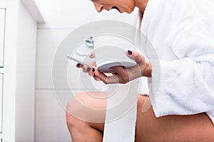 Woman sitting on toilet writing text message on cell phone