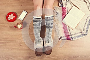 Woman sitting on the wooden floor with cup of coffee, phone, cookie and book. Close-up of female legs in warm socks with a deer vi